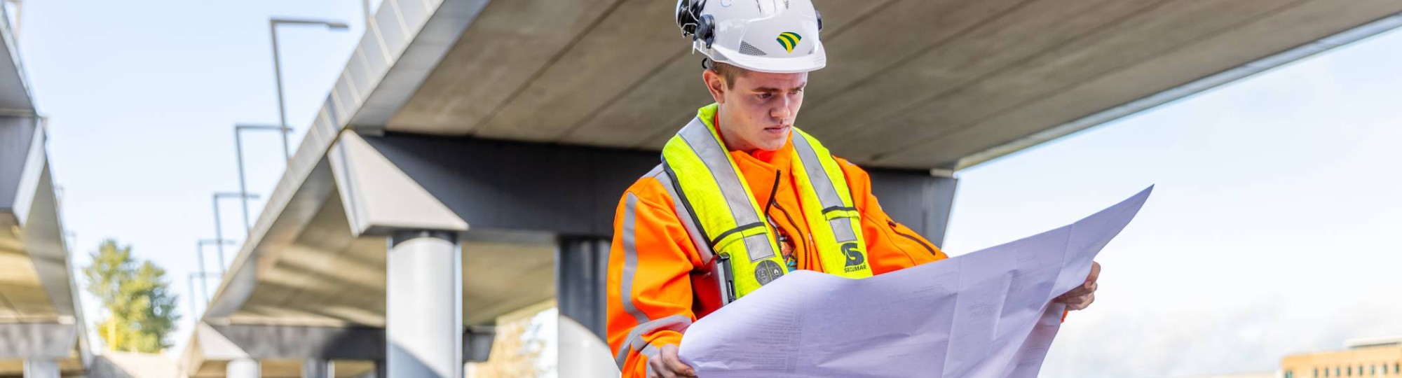 Man met veiligheidshelm en oranje werkjas checkt de bouwtekening onder een viaduct in aanbouw.