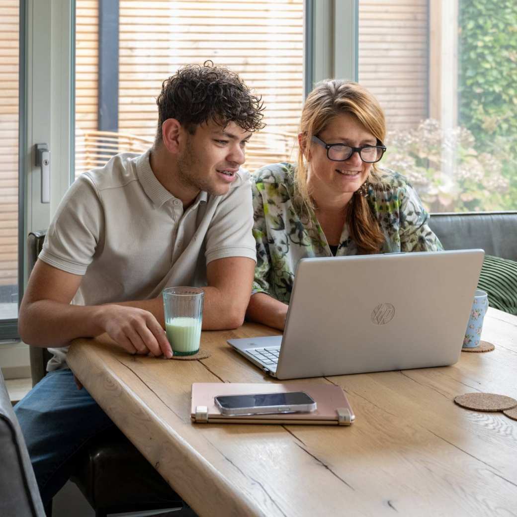 Moeder en zoon zitten aan een tafel en kijken lachend naar een laptop. 