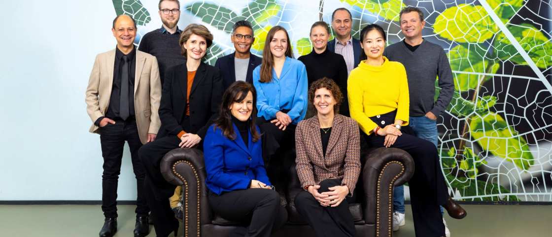 11 people standing in front of a wall with an image of mint leaves in front of a blue sky. 5 of them sit on a sofa and the other 6 stand around them. Everyone is facing the camera and smiling.