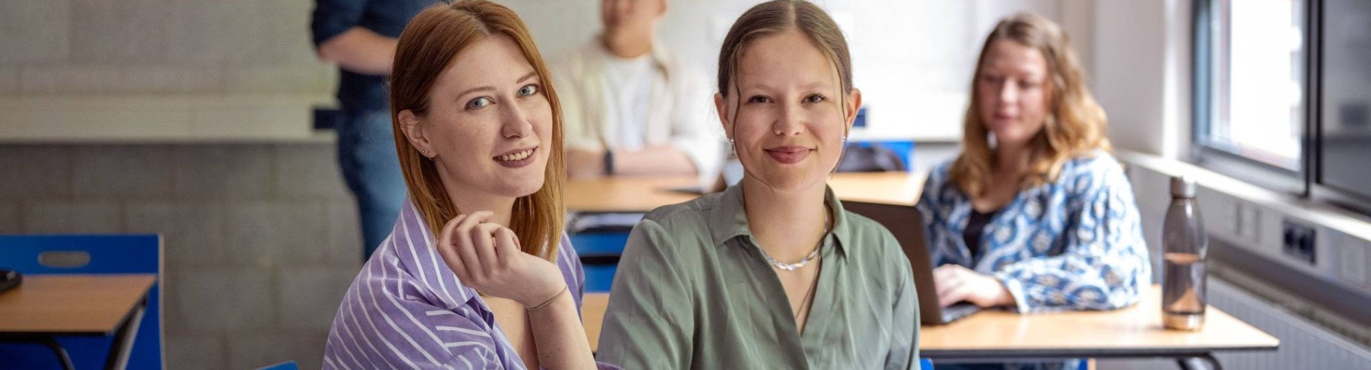 Twee studenten zijn samen aan het werk op de laptop, kijken in de camera, zijn in een klas.