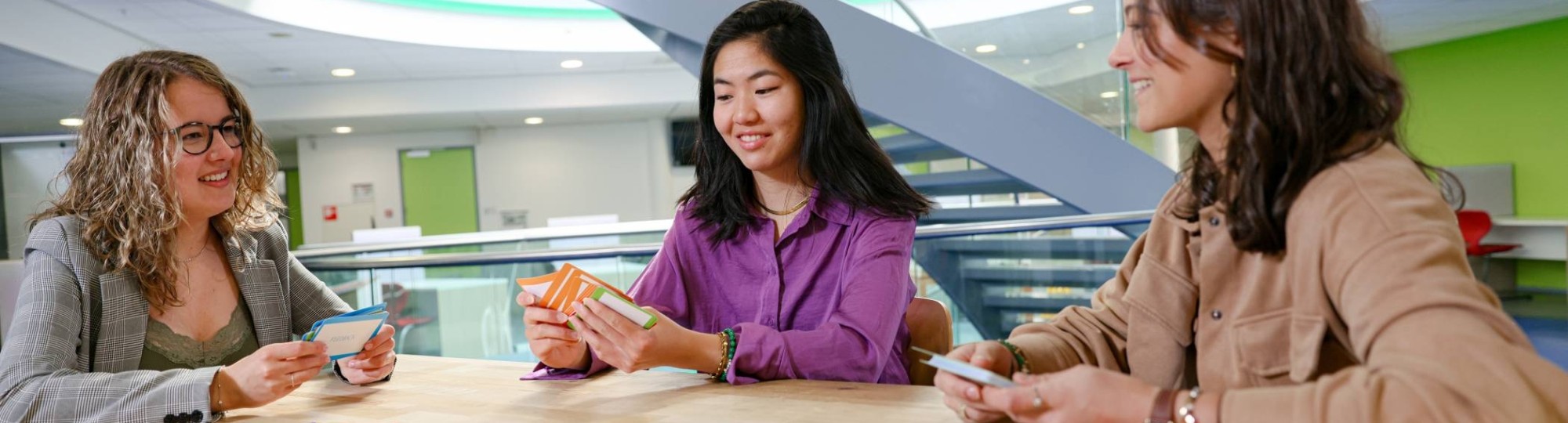 drie studenten van de opleiding toegepaste psychologie spelen een kaartspel aan een houten tafel