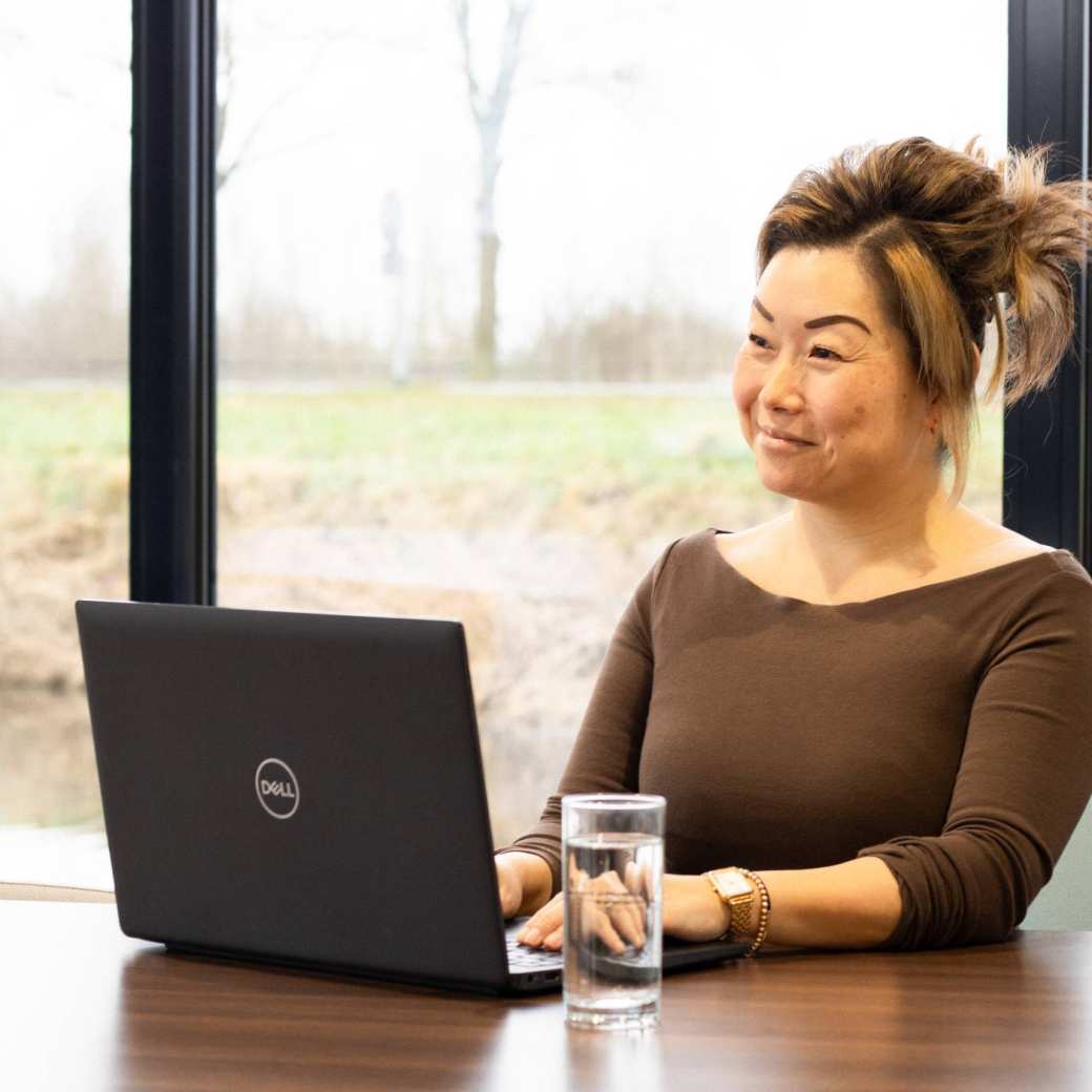 Dame poseert met een glimlach, achter een laptop. Een glas water staat naast haar op het bureau.