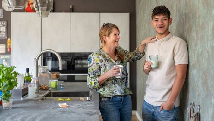 Moeder en zoon lachen en staan in de keuken, terwijl de moeder eeen hand op de schouder van haar zoon legt.