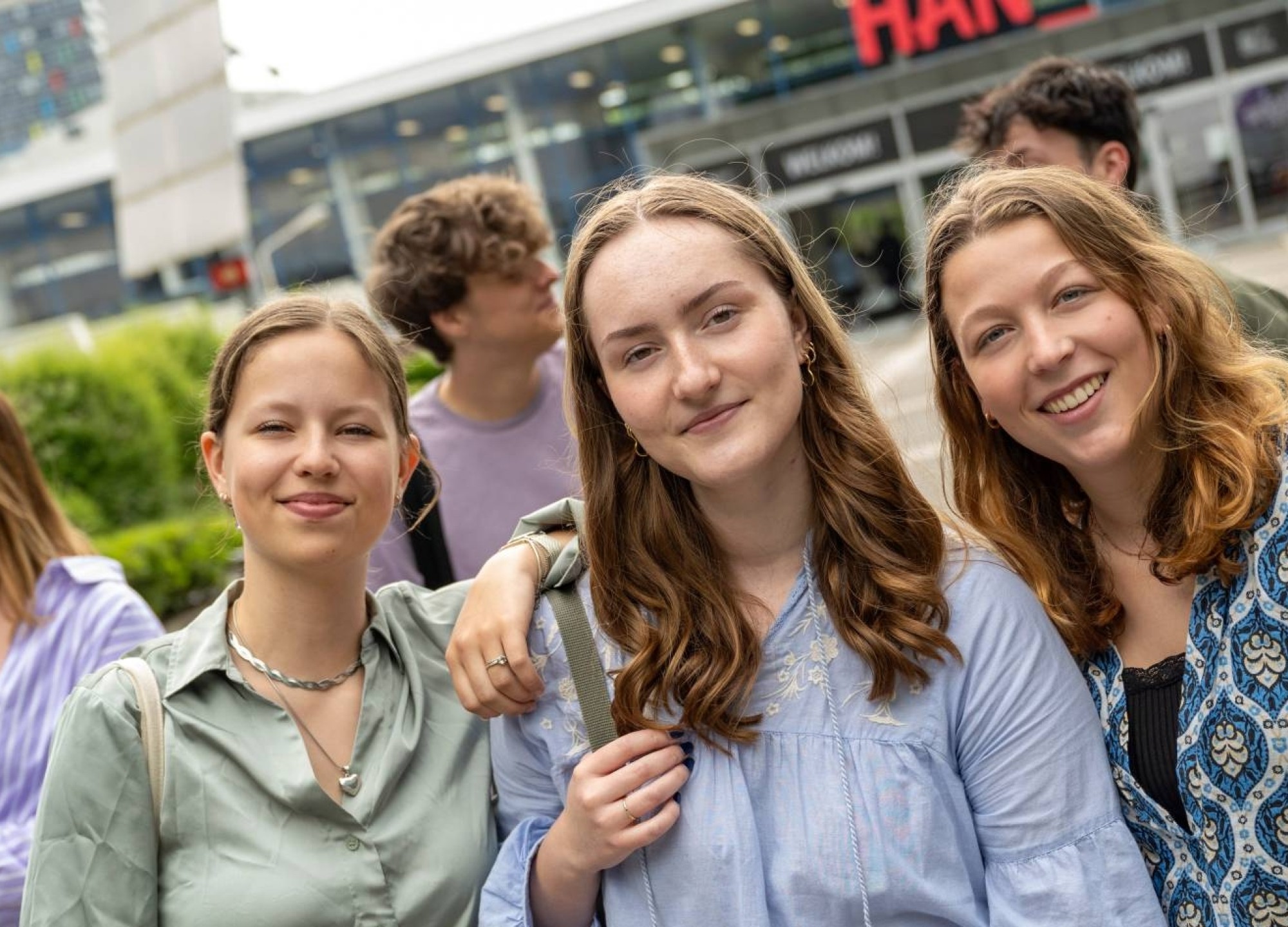 Studenten staan met vriendengroep buiten bij de ingang van gebouw R26, kijken in de camera.