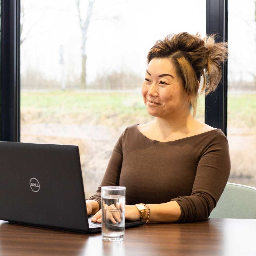 Dame poseert met een glimlach, achter een laptop. Een glas water staat naast haar op het bureau. 