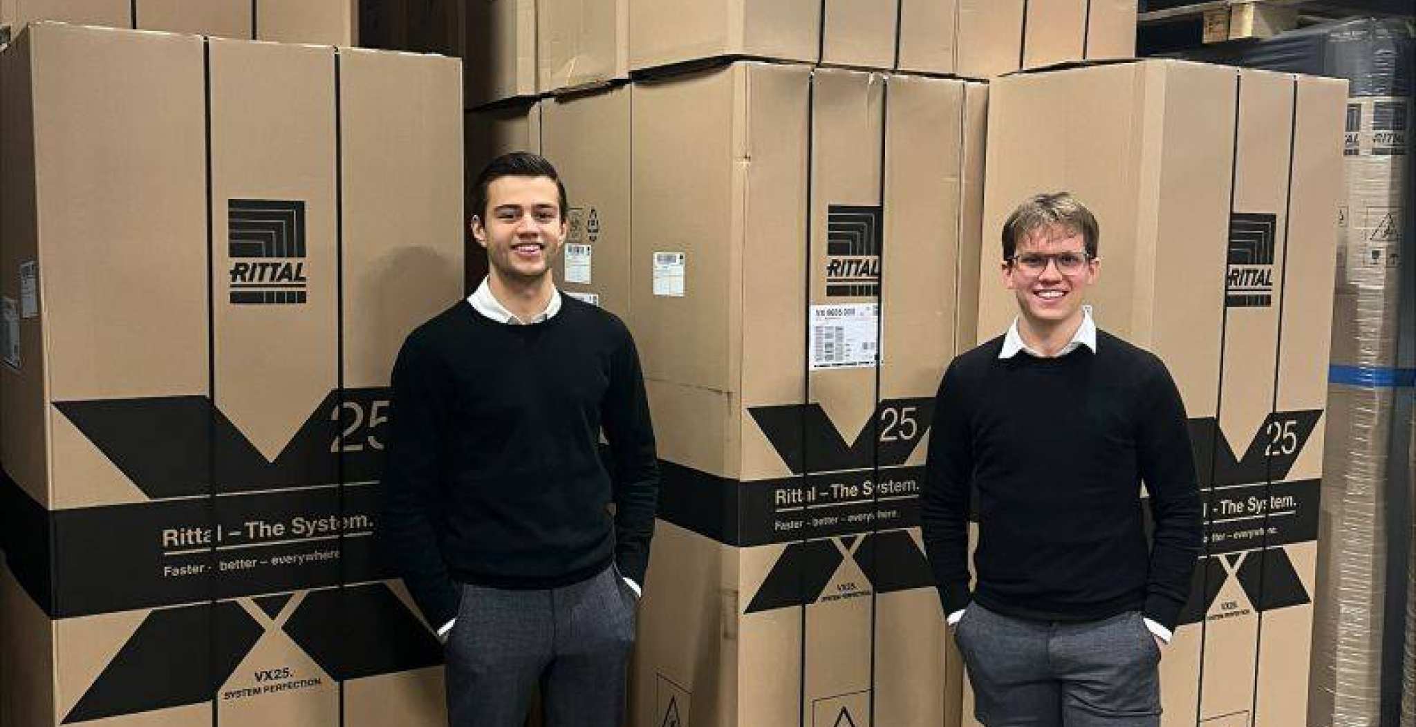 Quinten Stuurman en Guus Roos, studenten Technische Bedrijfskunde, staan in het magazijn van Rittal Nederland. Op de achtergrond zie je grote hoge dozen staan om vervoert te worden.