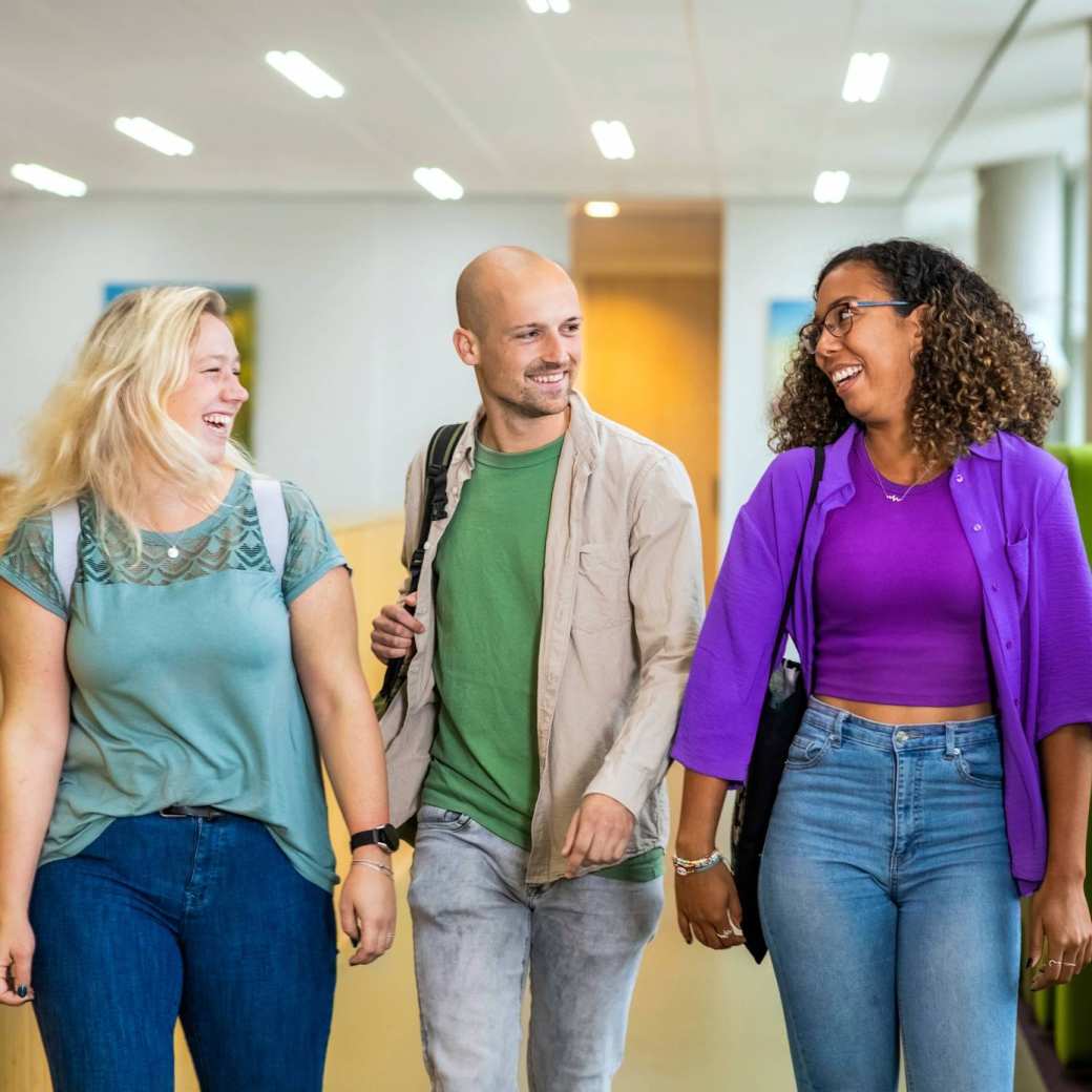Drie studenten Leraar Wiskunde lopen lachend naast elkaar op de gang in de school
