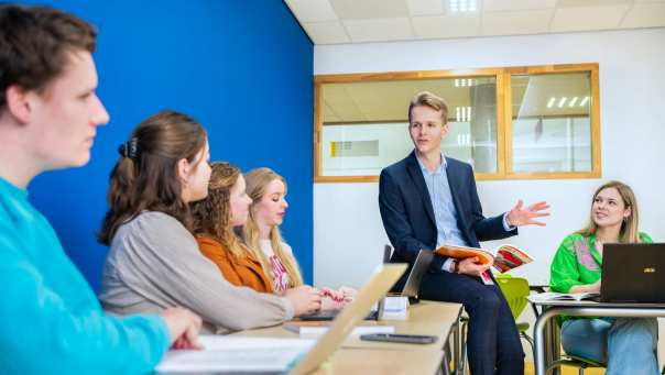 Docent Leraar Duits bladert door studieboek en praat tegen studenten in de klas.