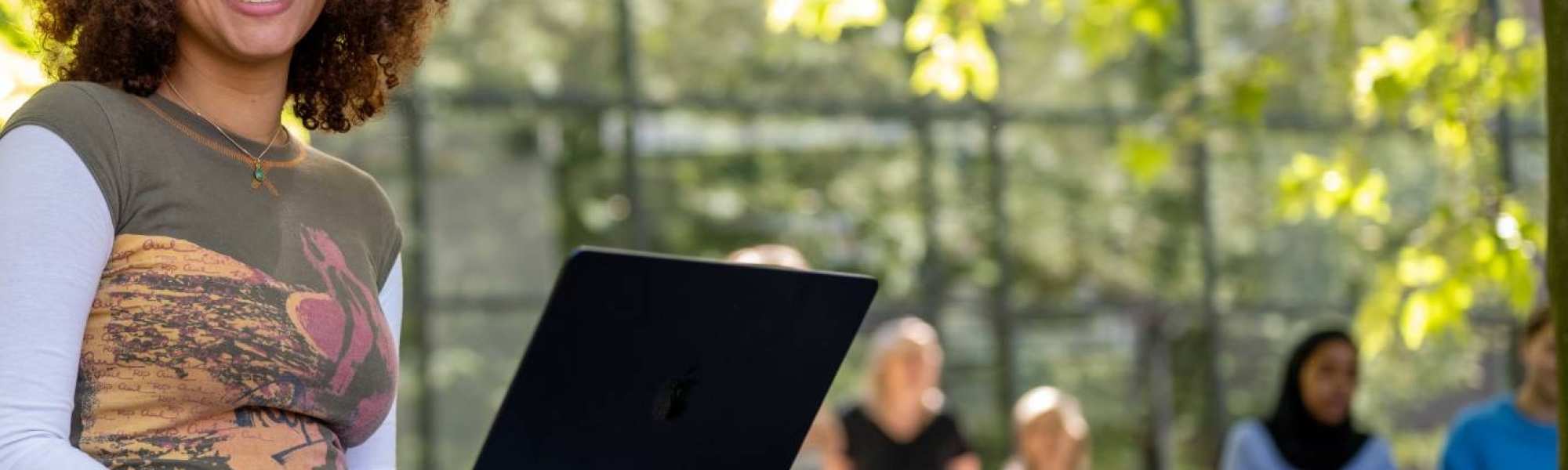 Studente kijkt in de camera, met laptop op schoot, zittend op de tafel, buiten bij K33. 
