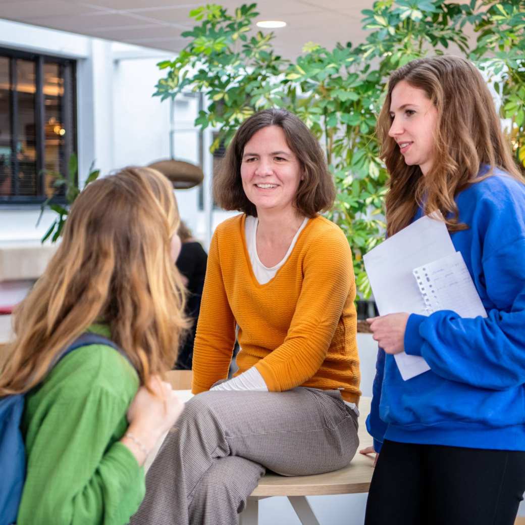 3 studenten gekleed in oranje, blauw en groen, overleggen met elkaar op de gang.