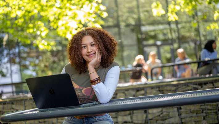 Studente kijkt in de camera, hand onder haar hoofd, werkt op laptop, buiten op bankje bij K33.