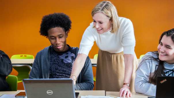 Docente Leraar Engels wijst lachend iets aan op de laptop van een student tijdens de les.