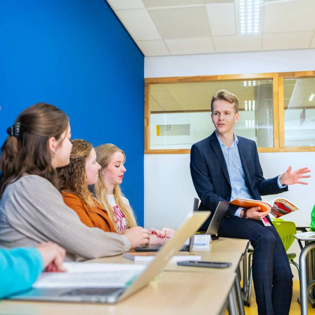 Docent in donkerblauw pak zit op de tafel met een boek in zijn hand, terwijl hij zijn studenten uitleg geeft