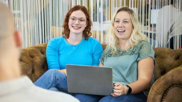 Twee studenten Leraar Wiskunde zitten naast elkaar met de laptop op schoot op de bank. Ze lachen naar de docent die tegenover hen zit.