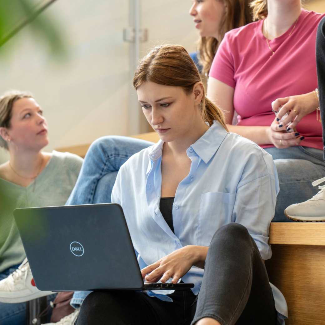 Studenten zittend op de trap in de hal van gebouw R26, werken op de laptop.