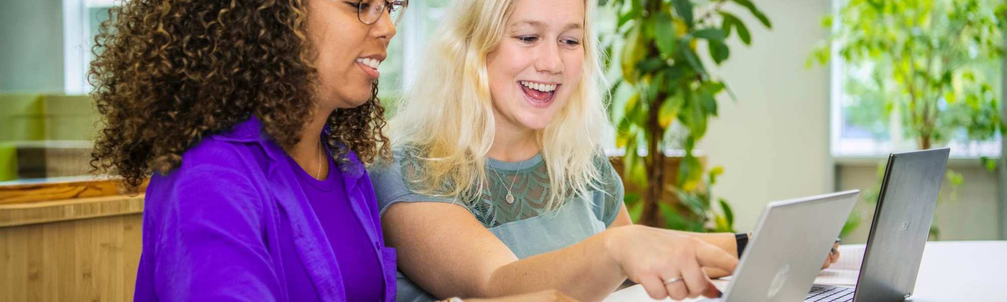 Twee studenten Leraar Wiskunde zitten naast elkaar met een laptop. De ene wijst lachend op het scherm iets aan en de ander kijkt lachend mee.
