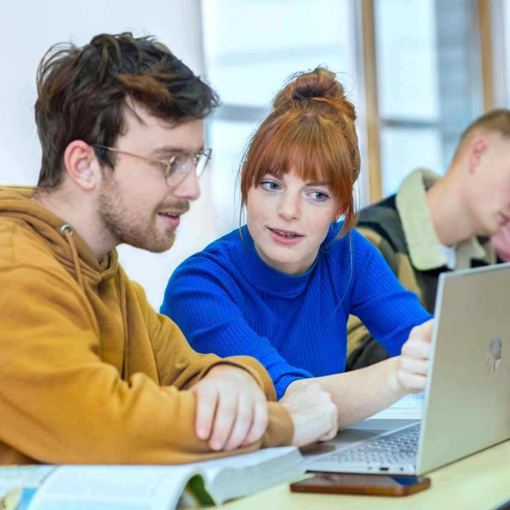 Twee studenten zitten naast elkaar en kijken samen aandachtig naar de laptop die voor hun staat.