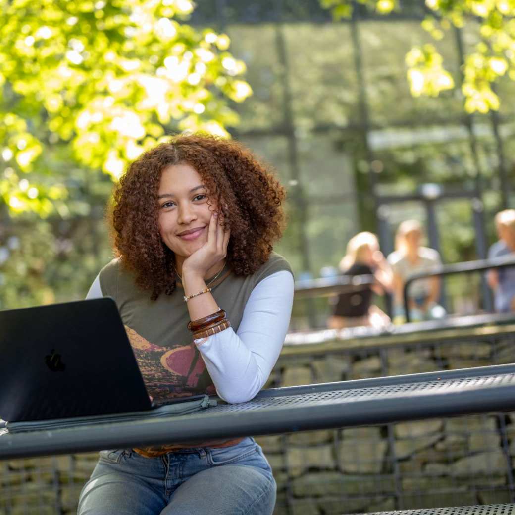 Studente kijkt in de camera, hand onder haar hoofd, werkt op laptop, buiten op bankje bij K33.