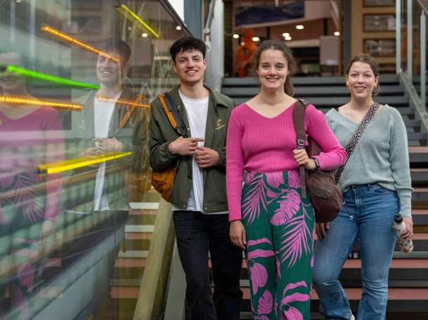 Drie studenten lachen in de camera, staan op de trap in lichtstraat van gebouw R26. 