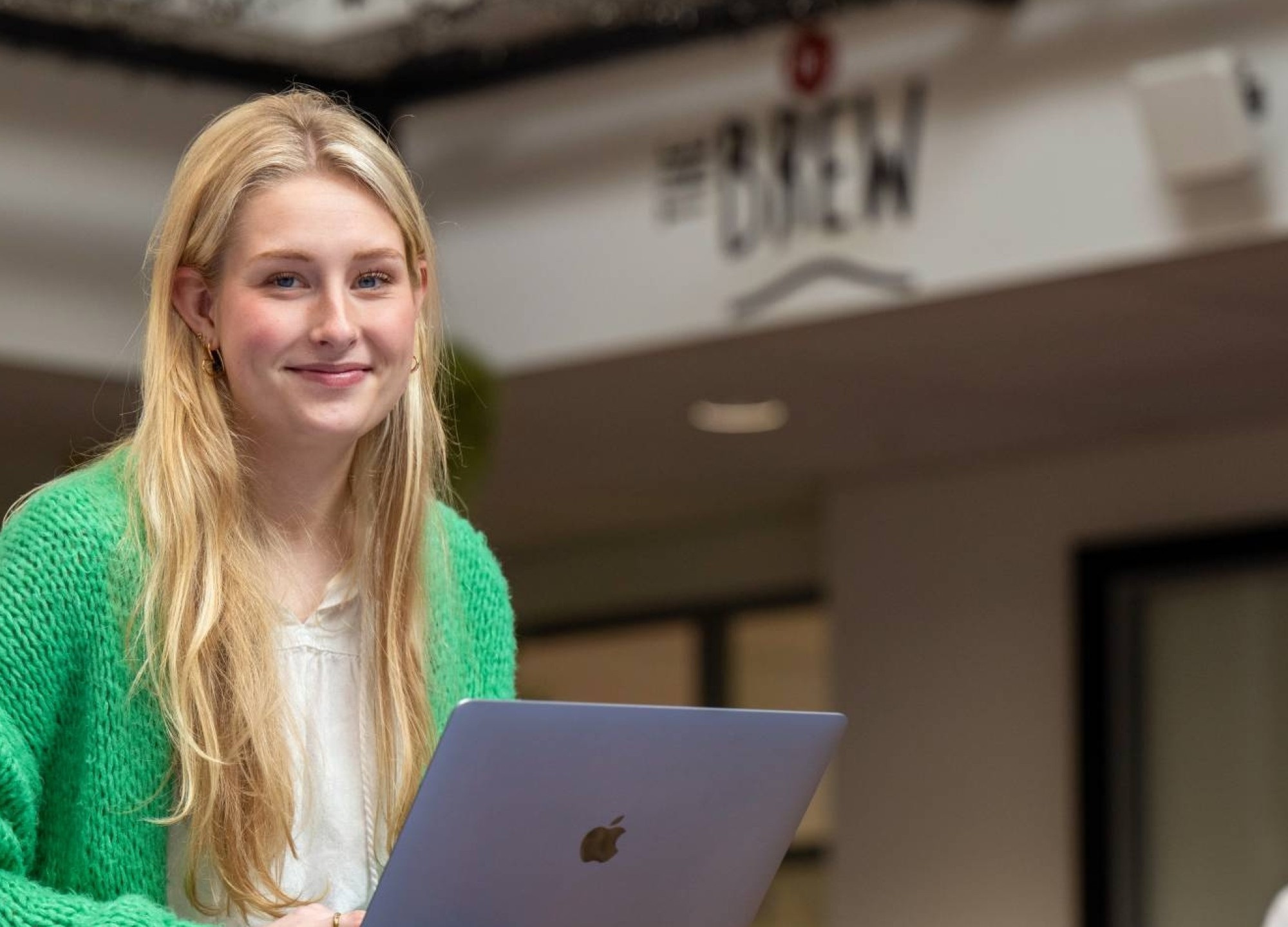 Studente lacht in de camera, heeft laptop op schoot en is aan het werk bij The Brew. 